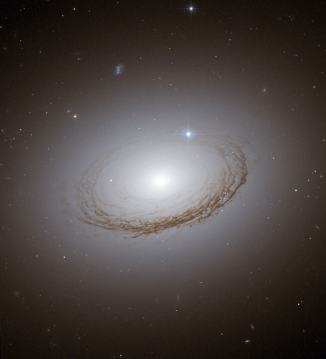 Hubble Space Telescope image of NGC 7049, a large galaxy with a bright glowing core surrounded by faintly glowing, smooth halo-like light. Dark dust lanes form ring patterns circling the center. Numerous distant stars and galaxies are scattered across the background.