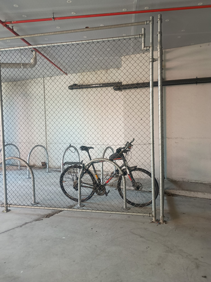 A bicycle sits lonely in a fenced off cage, locked at both ends to one of those floor hoop thingies.  In the background, a sinister black pipe emerges from the concrete wall and goes to places unknown.