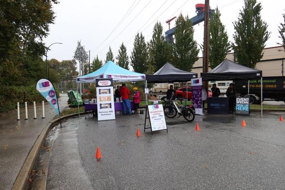 Station celebrating Go By Bike Week in North Vancouver. There are 3 tents and some signs. A bike is parked in front of one of the tents. The ground is wet from rain.