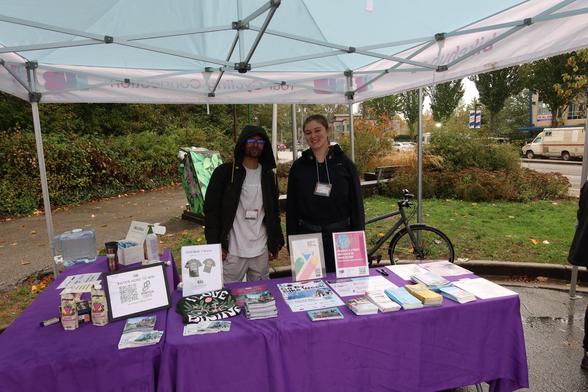 Two of the staff behind a table with some maps, other resources, and snacks. The table is under a tent and a bike is parked behind the tent.