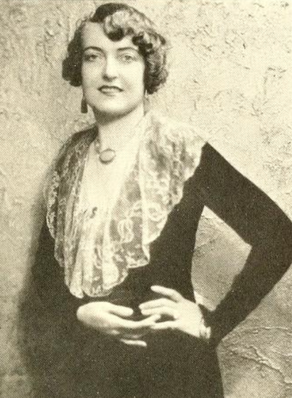 Mabel Sykes, about 1930, a smiling white woman, standing, wearing a  dark dress with a drapy lace collar; her hands are together on one hip