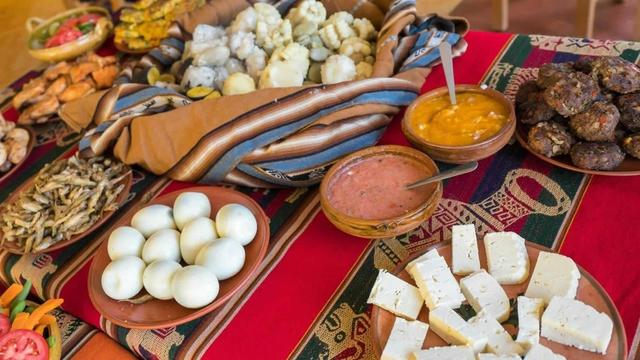 En la imagen se ve un apthapi con diversas comidas tradicionales sobre un aguayo, un mantel colorido. Hay papas, choclo, huevos y trozos de queso fresco, rodeados de cuencos con salsas de colores cálidos.