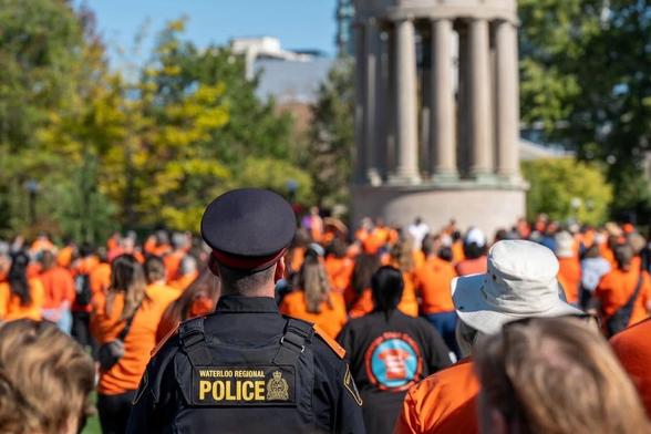 A police officer, wearing a uniform with "Waterloo Regional Police" visible, stands with their back to the camera, observing a large crowd of people in orange shirts gathered in Victoria Park, Kitchener.