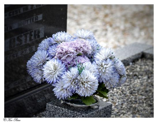 A close up photograph of a bouquet of artificial flowers with white and blue tipped petals surrounding a cluster of faded purple blooms.

The bouquet sits on a grey granite memorial base against a blurred background of light-coloured gravel. A darker granite headstone, partially visible on the left, features carved text.

The photo is watermarked '© Kev Peirce' and 'Gardens of the Silent'.