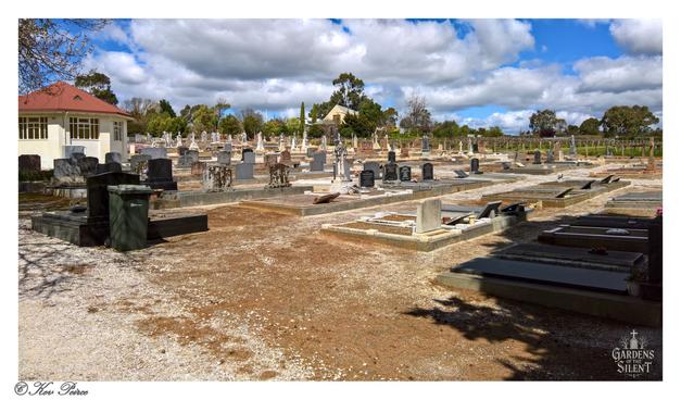 A panoramic view of St. John's Cemetery in Tanunda under a partly cloudy blue sky.

Rows of gravestones and flat grave markers stretch across a landscape of reddish-brown earth and light gravel paths.

In the foreground, shadows from unseen trees are cast on the ground. To the left, a light-coloured building with a red roof is visible.

In the distance, more trees and a small white building, possibly a church, can be seen, along with what appears to be a vineyard.

The image is watermarked '© Kev Peirce' and 'Gardens of the Silent'.
