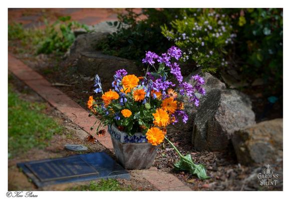A close up, shallow-depth-of-field photograph showing a ceramic pot of fresh flowers.

The bouquet is dominated by vibrant orange calendula or marigolds and tall sprigs of smaller purple wildflowers.

The pot sits on a brick and dirt path next to a dark, rectangular grave marker or plaque, which is blurred in the foreground.

Large stones and green foliage, including small white and purple flowers, frame the background.

The image is watermarked '© Kev Peirce' and 'Gardens of the Silent'.