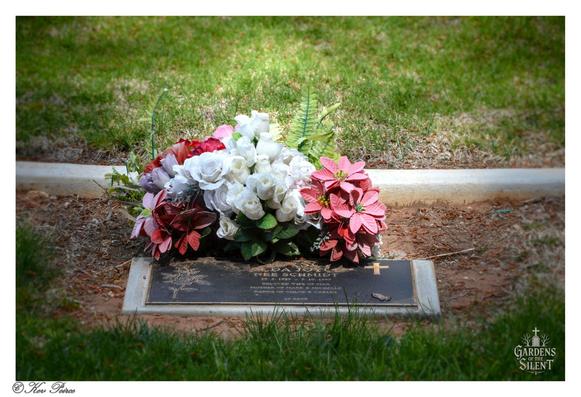 Ground level remembrance: White roses and pink poinsettias on a quiet, grassy plot.

A low angle, close-up photograph of a bronze grave plaque set into the grass.

Resting on the plaque is a large bouquet of artificial flowers, primarily white roses and striking deep pink poinsettia blooms, surrounded by green foliage.

The plaque, which includes a cross and a butterfly motif, is set against a background of green and brown earth, with a concrete curb visible behind it. The foreground is framed by blades of grass.

The image is watermarked '© Kev Peirce' and 'Gardens of the Silent'.