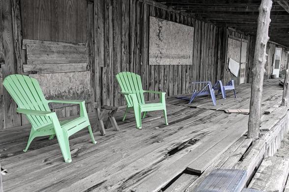 A landscape photo where the background, which is the covered porch on a long abandoned frontier wood planked commercial building, is in a nearly black and white rendering and the four modernish plastic chairs on the porch are brightly colored. The view is looking down the porch from the lower left toward center right. There's a rough round pole on the far right that holds up that section of the porch roof. The wall of the building is vertical wood planks with boarded up windows and a door. The first two chairs on the left, and which face outward toward the right are a bright yellow-green. Between them sits a small half broken table. The last two chair further down the porch are purple and the backs are broken off.
