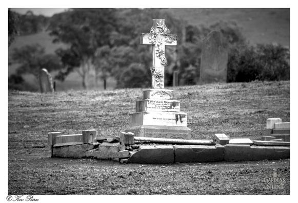 A black and white photo of a white, ornately carved stone cross, decorated with floral reliefs, stands atop a square pedestal and base in a dry, grassy cemetery.

The cross is slightly weathered and appears to be the central monument of a grave, surrounded by a low, partially collapsed, stone border in the foreground.

Several other weathered headstones, one of which is a dark, rounded slab, are partially visible in the blurred background.

Rolling hills with scattered gum trees are visible further in the distance under a pale sky.