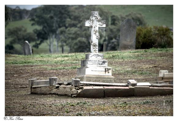 A white, ornately carved stone cross, decorated with floral reliefs, stands atop a square pedestal and base in a dry, grassy cemetery.

The cross is slightly weathered and appears to be the central monument of a grave, surrounded by a low, partially collapsed, stone border in the foreground.

Several other weathered headstones, one of which is a dark, rounded slab, are partially visible in the blurred background.

Rolling hills with scattered gum trees are visible further in the distance under a pale sky.