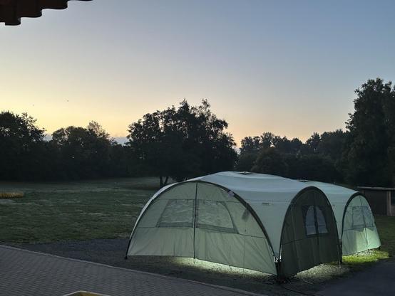 Ein großes, hellgrünes Zelt steht auf einer Wiese am Rand eines gepflasterten Platzes. Es ist früh am Morgen oder am Abend, der Himmel zeigt ein sanftes Farbspiel von Blau zu Orange. Hinter dem Zelt stehen hohe Bäume im Dämmerlicht. Aus dem Zelt scheint Licht nach außen, sodass es von innen beleuchtet wirkt.