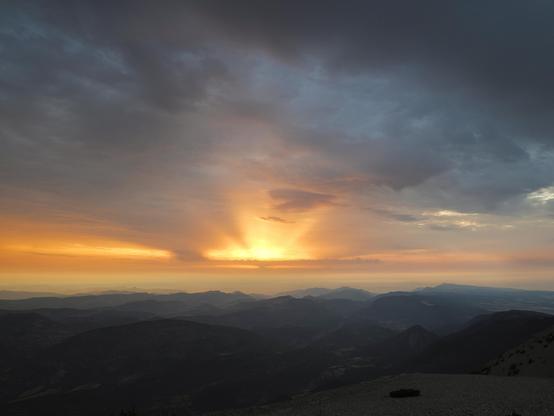 A scenic view of a sunrise over mountainous terrain, featuring dramatic clouds with a gradient of orange, pink, and blue hues in the sky. The silhouette of rolling hills and valleys is visible in the foreground.