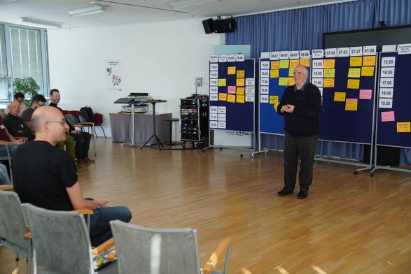 The Open Space Marketplace. The facilitator stands in front of multiple pin boards which show the program of the day. Some people from the audience can be seen on the left of the picture.