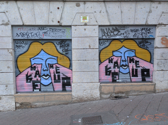 Sur deux rideaux métalliques fermés sur des vitrines carrées dans un mur de pierre dans une rue en pente, deux tags identiques représentant un visage bleu, encadré par des cheveux blonds mi-longs, la bouche maquillée, les yeux fermés d'où sortent deux rayons larges et roses où il est écrit en lettres capitales noires dispersées "Wake Me Up".