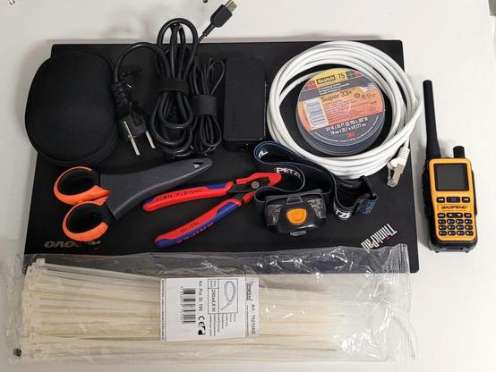 Auf dem Bild liegt eine Auswahl an Werkzeugen, Kabeln und Geräten ordentlich nebeneinander. Alles ist auf einer schwarzen Lenovo-ThinkPad-Laptopunterlage platziert.

Von links nach rechts sieht man:

Ein kleines schwarzes Etui.

Ein Netzkabel mit Eurostecker und ein weiteres Kabel, zusammengerollt mit Klettverschluss.

Ein schwarzes Netzteil.

Eine Rolle Isolierband (Marke Scotch Super 33+).

Ein zusammengerolltes weißes Netzwerkkabel (LAN-Kabel mit RJ45-Stecker).

Eine Schere mit orangefarbenen Griffen.

Eine rote und blaue Zange (Seitenschneider).

Eine schwarze Stirnlampe mit orangener Taste (Marke Petzl).

Rechts daneben ein gelbes Handfunkgerät mit Antenne (Marke Baofeng).

Unten, quer über das Bild gelegt, ein großes Bündel weißer Kabelbinder in Plastikverpackung.


Die Gegenstände wirken wie eine kleine Ausrüstung für Reparatur-, Netzwerk- oder Outdoor-Einsätze.