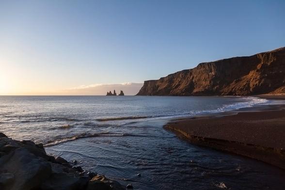 Standing on a jetty formed of large, dark boulders on a beach at the boundary where the water meets the sand, the view is partly out to sea, but mostly focused on the cliffs that come from the right side of view down towards the horizon, abruptly terminating in the centre of the image. The cliffs, like the sand on the beach that is just visible as a crescent curving away from near the viewpoint around and towards the cliffs, are dark rock. While the sky is mostly clear, except for a band of cloud on the horizon, the sun is obviously low in the sky and weak, lending everything little warmth and leaving the water near the viewer almost black. Seemingly on the horizon, although actually closer than that, out to sea a little way from the end of the cliffs, are jagged rocks jutting up, like the silhouettes of stalagmites or chipped teeth.