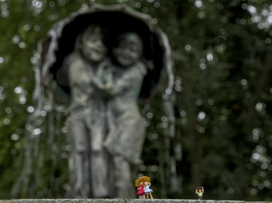 vor einer brunnen-bronce (zwei kinder unter einem regenschirm) stehen an der kannte miniaturfiguren. einmal zwei mädchen unter einem regenschirm, einmal ein junge, der einen koffer über den kopf hält