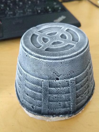 A very concrete-looking piece of terrain, shaped like a tapered cone, with industrial looking ridges on its side. It's been painted black and then drybrushed in white. The mild it was made from was originally a Ricotta packaging.