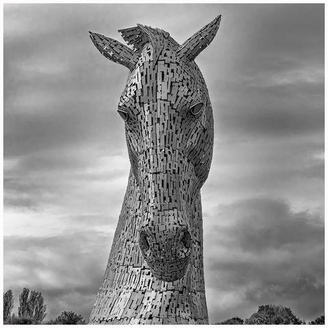 A large, abstract sculpture of a horse's head, constructed from irregularly shaped metal plates. The background features a cloudy sky.