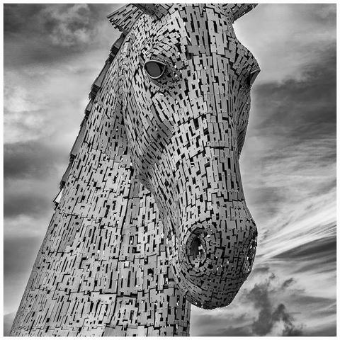 A large, abstract sculpture of a horse's head, made from interlocking metal pieces, against a dramatic sky. The image is presented in black and white.
