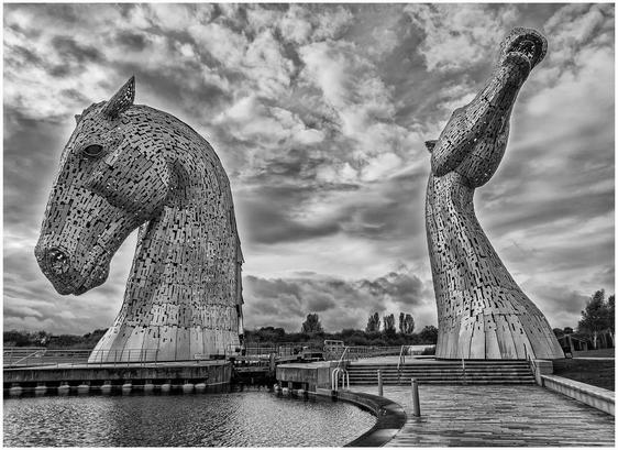 The image features two large, abstract sculptures of horse heads, crafted from metal plates, situated near a body of water. The scene is in black and white, showcasing a dramatic sky with clouds, and the sculptures are set against a tranquil landscape.