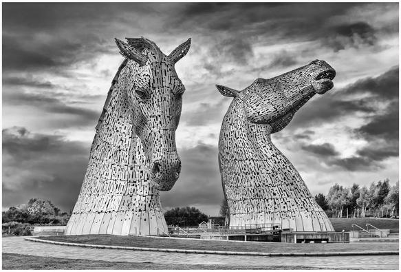 A black and white photo of two large horse-head sculptures, made from intricately arranged metallic pieces, set against a dramatic sky. The surroundings include pathways and greenery.