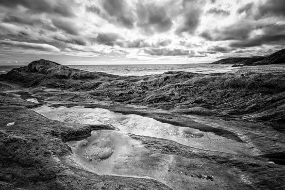 A dramatic black and white photograph of a rugged, rocky coastline under a powerful, cloudy sky. The foreground shows textured dark rocks with a small, reflective pool of water creating a slight highlight. The horizon features a dark, calm sea meeting the distant landmass on the right. The overall mood is intense and moody.