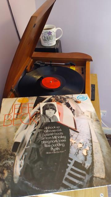 Record player with a 12" black vinyl record.
In the foreground is an album cover with a small black and white photo of Jeff Beck over a close up colour photo of the front wheel of a motorbike parked on a kerbside drain.
Behind the player is a mug of coffee on a speaker.