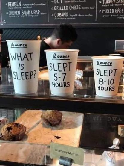 Photo of a coffeehouse and bakery. Three paper cups of different sizes are standing on top of the glass baked goods display, each with writing on it. The smallest cup reads "Slept eight to ten hours." The middle cup reads, "Slept five to seven hours." The largest cup reads, "What is sleep?"