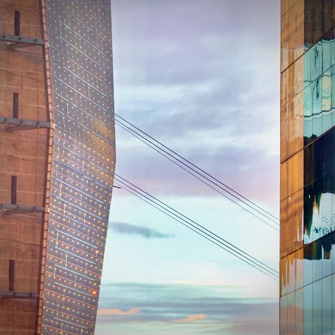 The sides of two glass-facade buildings face each other, seemingly connected via two groups of three diagonal power lines against an orange and blue, partly cloudy morning sky.