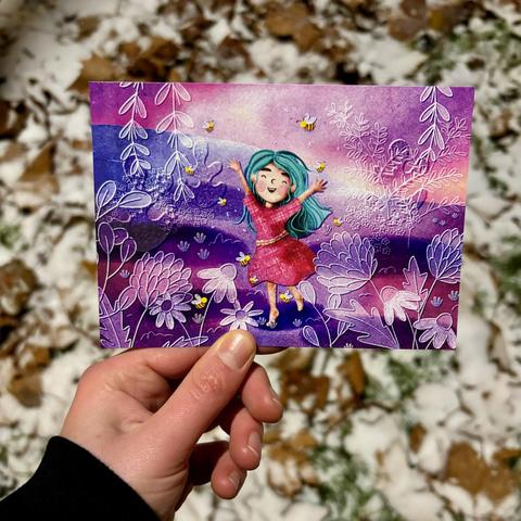 Foto von einer Postkarte. Die Postkarte zeigt die Illustration von einer Person mit blauen Armen in einem roten Kleid, die die Arme freudig in die Luft reckt. Die Person ist umgeben von Bienen. Überall befinden sich Blumen in weißem Linework auf rot-violettem Untergrund.