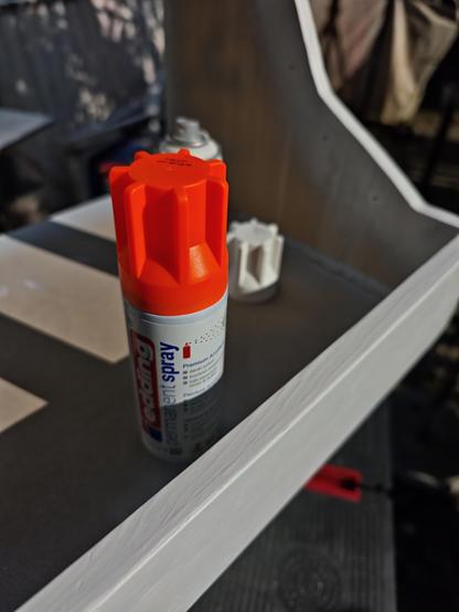 A crisp close-up of an Edding spray can in bright neon orange, standing upright inside a freshly primed Eurorack studio case. The matte wood surface underneath shows a two-tone paint job, part white and part raw MDF, illuminated by sharp afternoon sunlight. The can's star-shaped cap glows with intense orange energy, casting hard shadows across the frame. In the background, another can peeks in with a white cap, and next to it lies a detached spray nozzle cap from the white paint can, shaped like an oversized gear. The entire shot radiates focused maker vibes under a clear autumn sky.