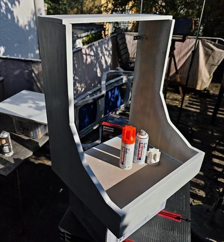 An outdoor workbench scene featuring a Eurorack studio case mid-build, standing tall and proud in blazing afternoon light. The case is partly painted in cool white and grey, with clean edges and primer streaks still visible. Resting inside the lower tray are two Edding spray cans... one uncapped, revealing its vibrant neon orange lid...  and the gear-like cap from a white spray can. Tools and paint cans scatter the background, with dappled tree shadows dancing across the work area. The whole setup screams DIY ambition with a hint of precision chaos... the kind only a synth-head in the zone can love.