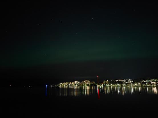 Green northern lights above a lake and city lights. The lake reflects the city lights. Above the northern lights the Big Dipper constellation is visible.