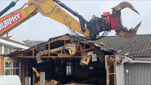 Screenshot shows an excavator with its ripper arm lifted in the air, poised to continue tearing down Cwmfelin Social Club in Cwmbwrla, Swansea.