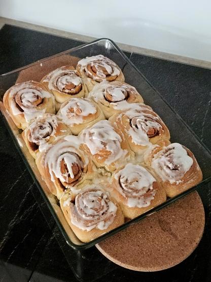 Foto einer gläsernen Auflaufform mit 12 Zimtschnecken mit Zuckerguss darin.