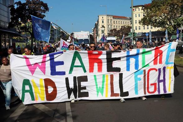 Das Foto zeigt die Demonstration mit Transparent, auf dem der Text „We are here and we will fight“ in bunten Buchstaben steht.