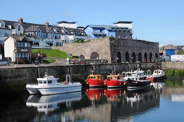 The harbour at Seahouses in Northumberland. The image shows a view over a harbour to a quayside extending in from the left with small fishing boats moored to it. Above the far end of the quayside is a row of stone storage sheds with brown doors. Behind and above them are other buildings including a white-painted hotel. The sky is blue.