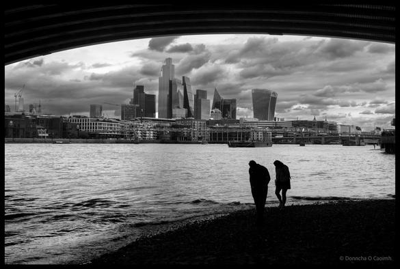 Black and white photograph framed by curved bridge arch of Blackfriars Railway Bridge showing silhouetted couple standing at Thames shoreline with dramatic cloudy sky above London's modern skyline, including the Leadenhall Building, Cheesegrater, and other City skyscrapers across the river.