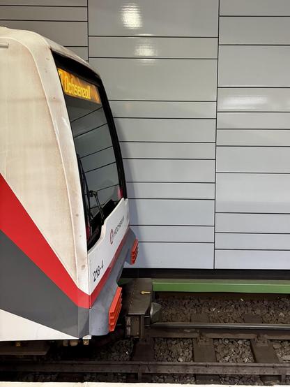 Eine Nahaufnahme der Vorderseite eines Zuges mit einem Display, das das Ziel "Ochsenzell" zeigt. Der Zug befindet sich in der Nähe einer modernen Bahnhofswand mit horizontalen Paneelen. Das Gebiet umfasst auch sichtbare Zuggleise und Kies.