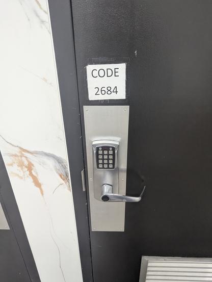 The image depicts a close-up view of a door with a keypad lock. The door is predominantly black with a textured surface. To the left of the door, a section of a wall is visible, featuring large white tiles with a marble-like pattern of brown and gray veins.

The main focus is the silver keypad lock, which is mounted on a rectangular, silver plate. The keypad has a standard numerical layout with a few additional function buttons. A silver handle extends from the lock, curving outward. Above the lock, a small, rectangular piece of paper is taped to the door. This paper reads "CODE" followed by the number "2684".

The composition is straightforward, with the door dominating the frame. The lighting appears to be even, highlighting the textures of the door and the metallic sheen of the lock. The overall impression is one of security and access control.