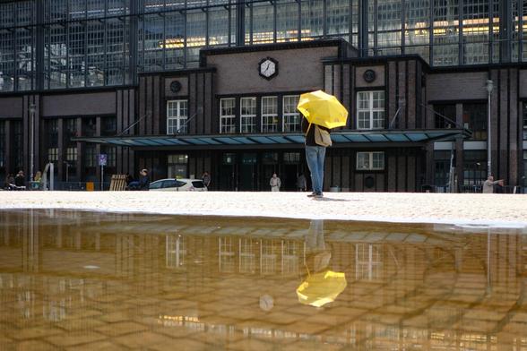 Das Bild zeigt ein großes, architektonisch interessantes Gebäude, wahrscheinlich ein Bahnhof oder ein ähnliches öffentliches Gebäude, mit einer Uhr an der Fassade. Im Vordergrund befindet sich eine Wasserpfütze, die das Gebäude und eine Person mit einem gelben Regenschirm reflektiert. Die Person steht vor dem Gebäude und hält den Regenschirm über sich. Im Hintergrund sind weitere Menschen und Auto zu sehen, was darauf hindeutet, dass es sich um einen belebten Ort handelt. The picture shows a large, architecturally interesting building, probably a train station or similar public building, with a clock on the facade. In the foreground is a puddle of water reflecting the building and a person with a yellow umbrella. The person is standing in front of the building, holding the umbrella over their head. In the background, other people and cars can be seen, suggesting that this is a busy place.