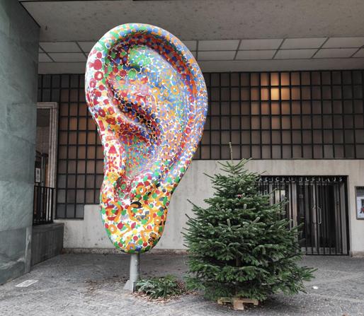 Large, brightly colored sculpture of a human ear. To the right of the sculpture, there is a small evergreen tree.