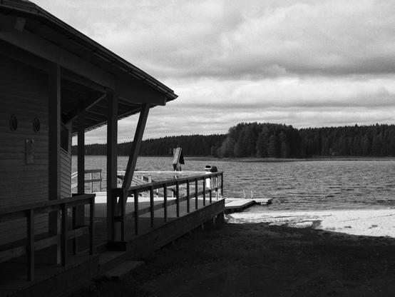 Black and white photo of early autumn lakeside scene with a sandy beach, a wooden pavillion on the left and dock extending into the calm water in the centre. The shoreline on the opposing side is forested, and a small forested island can be seen in the middle. The sky is cloudy.