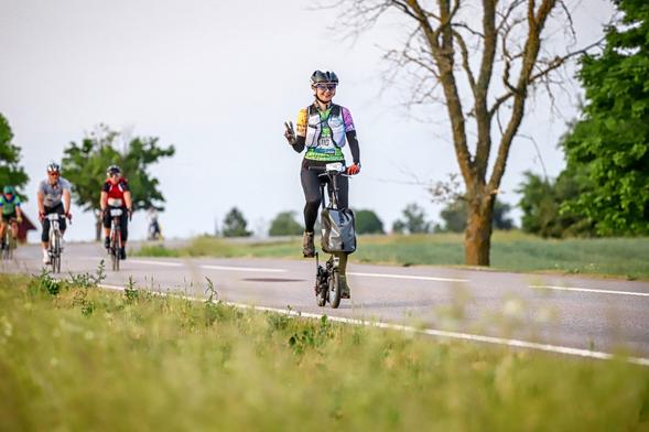 Person mit Startnummer auf einem Kwiggle mit 12" Reifen in aufrechter Haltung radfahrend vor sommerlicher weiter Wiesenlandschaft. Links im Bildhintergrund eine sich nähernde Rennfahrergruppe.