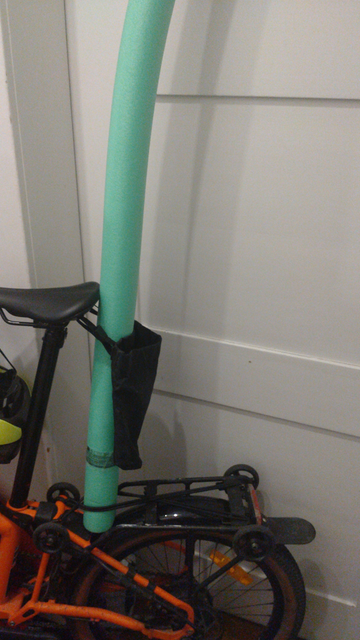 Eine grüne Schwimmnudel steht vor dem Gepäckträger eines orangefarbenen Brompton Electric G-Lines auf dessen hinterem Schutzblech und Controller, unten eingeklemmt zwischen zwei doppelt gekreuzten Gummistrapsen und oben zwischen Sattel und Satteltasche. Das G-Line ist an eine weiße Zimmertüre gelehnt.