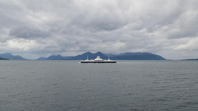 A photo of a large black and white ferry crossing a body of water with mountains in the distance. The sky is filled with clouds.