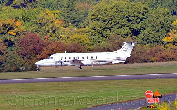 Slightly over-exposed side view of a twin propellor engined airliner taxiing from right to left.
The plane is white, with a thin black and gold stripe running along the body, and the golden registration "D-COCA" on the lower rear fuselage.
Green grass fills the foreground, lining both sides of a black runway.
Trees fill the background, with a grey house barely visible through the foliage.