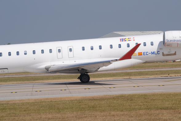Close-up of the red up-turned wingtip of a twin engined jet airliner taxiing from right to left.
The plane is almost entirely white, with a stylised yellow crown and a small Spanish flag next to the blue registration "EC-MLC"  on the lower rear fuselage, between the rear of the wing root and the white engine pods mounted on the upper rear fuselage.
There are blue "Scandinavian" titles on the sides of the engine pods.
Grass lines the sides of the grey runway, with the roof of a grey hangar visible over the top of the fuselage in the background, under blue-grey sky.