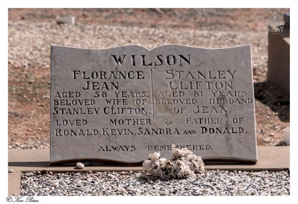 A modern, light coloured marble headstone for the Wilson family, located at Berri Cemetery.  The stone is carved in a scroll-top style and is dedicated to FLORANCE JEAN (Aged 58 years, Beloved Wife of Stanley Clifton) and STANLEY CLIFTON (Aged 81 years, Beloved Husband of Jean).  The bottom inscription reads: "LOVED MOTHER & FATHER OF RONALD, KEVIN, SANDRA and DONALD. ALWAYS REMEMBERED."  A small cluster of dried white or pale flowers rests on the light-colored gravel in front of the stone.